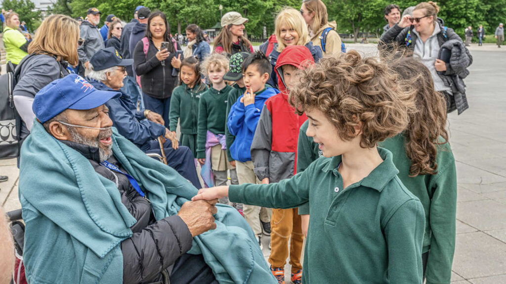 Group of kids and adults greet an elderly man in a wheelchair outdoors, shaking his hand.