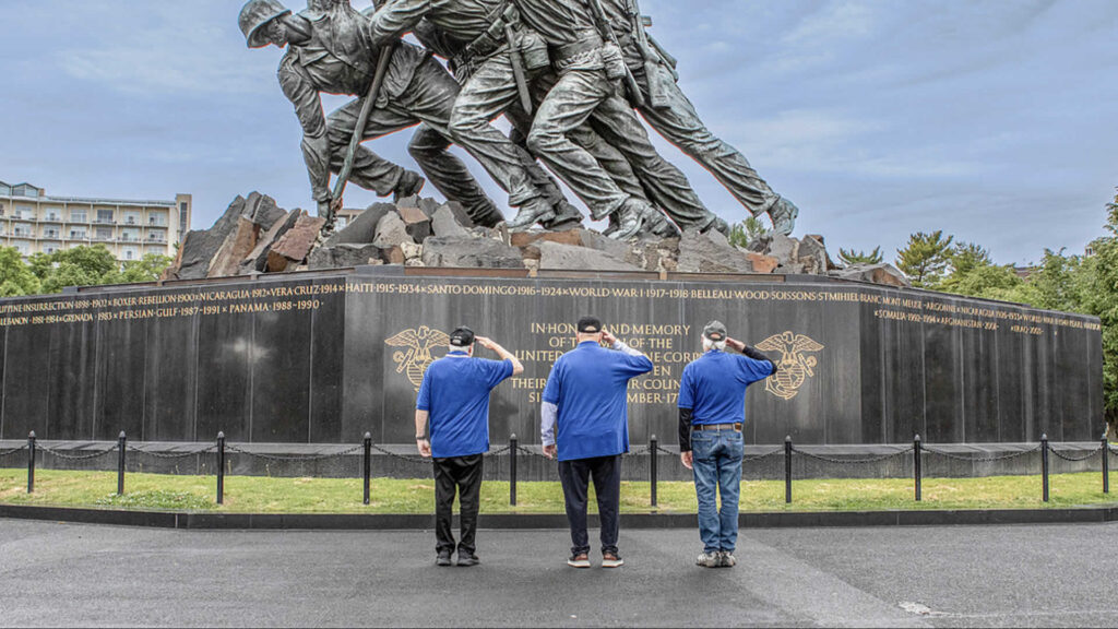 Three men in blue shirts salute a large war memorial with a dramatic sculpture of marching soldiers atop rocks behind a black wall with gold inscriptions.